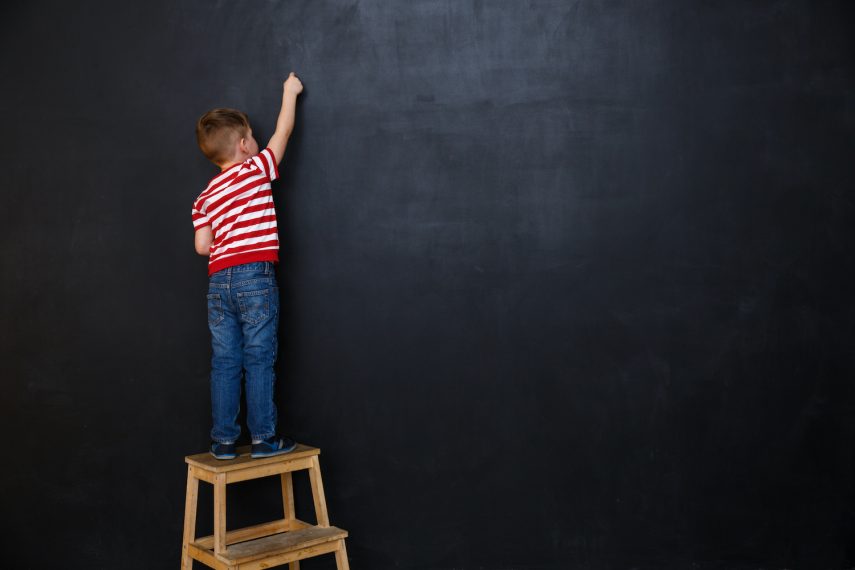 Back view of a little boy standing on ladder and writing with chalk on the backboard in school class